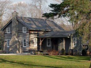 A log cabin is pictured; the wood of the cabin is greyed, and a stone chimney is shown to the left. The grass is green while surrounding trees suggest fall is nearing.