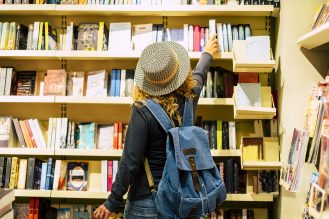 The back of a person wearing casual clothes, a hat and a denim backpack reaching up for a book from a full bookshelf in a library.