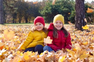 Two small children smiling, wearing coordinated yellow and red jackets and hats playing in the fall leaves