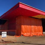 Construction site with the corner of a building covered in orange tarp and an unfinished red roof.