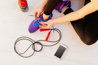 Top view of woman sitting on white wooden floor and tying her sport shoes. Skipping rope, bottle of water and mobile phone on the white wooden background. Sport, diet and healthy life concept.