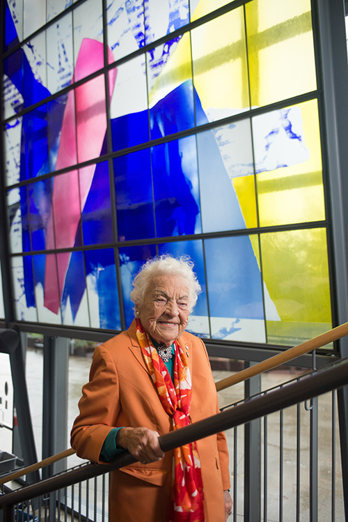 Hazel McCallion, former Mississauga Mayor, standing on a staircase with large panels of stained glass artwork in the background.