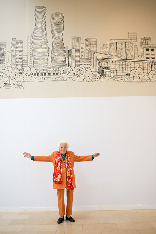 Hazel McCallion, former Mississauga Mayor, posing with her arms outstretched below a wall mural of the Mississauga downtown core.
