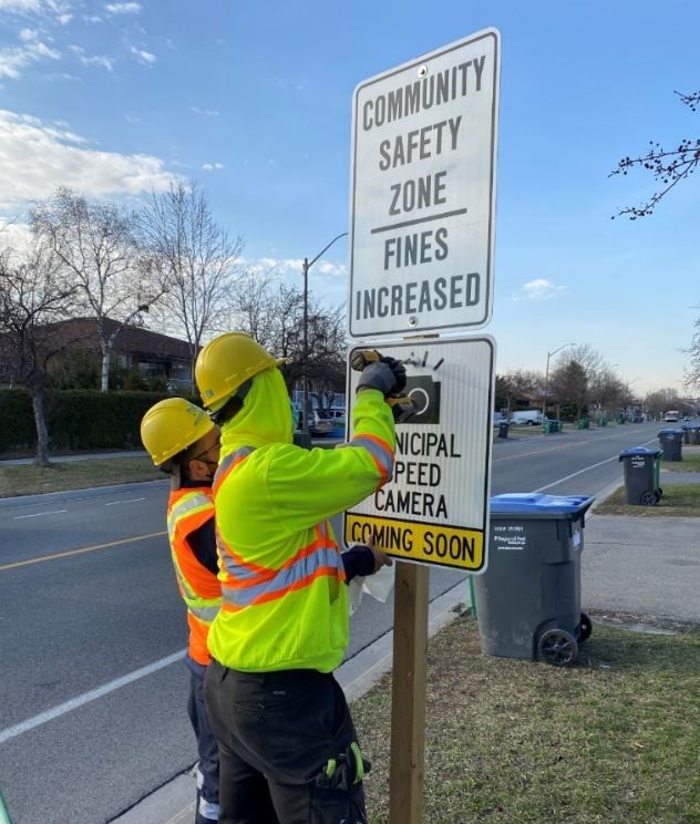 Two City of Mississauga staff in high-vis jackets installing an automated speed camera sign on a wooden post below a community safety zone sign