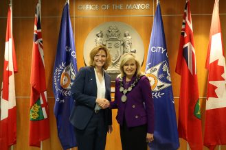 City of Mississauga Mayor & Dr Janet Morrison of Sheridan College shaking hands with flags & City emblem in the background