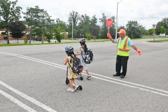 Two students walking their scooter across crosswalk with a crossing guard to get to school.
