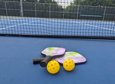 Pickleball equipment on the court including two racquets and two yellow outdoor pickleball balls