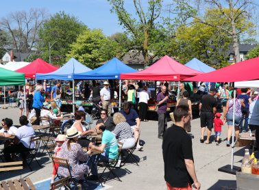 People sitting at picnic tables eating with vendors under pink and blue tents in the background