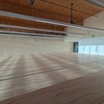 The fitness studio has hanging lights along the length of the ceiling and natural light on an uninterrupted floor.