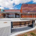 Wide, straight-on view of Carmen Corbasson Community Centre showing the full front entrance, the building's name signage, and long benches parallel to the building placed at a distance.