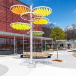Diagonal view of part of Carmen Corbasson Community Centre with a tall post featuring six circular fixtures and handing orange, yellow and green panels. The area includes benches, two trees and space for people to gather.