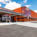 Diagonal view of Carmen Corbasson Community Centre entrace, showing a distinct light grey pedestrian pathway between dark asphalt for cars. The building features orange vertical panels and large glass windows.