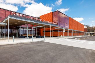 Diagonal view of Carmen Corbasson Community Centre entrace, showing a distinct light grey pedestrian pathway between dark asphalt for cars. The building features orange vertical panels and large glass windows.
