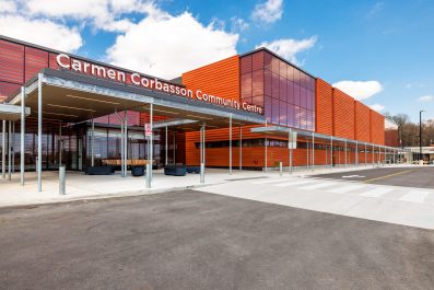 Diagonal view of Carmen Corbasson Community Centre entrace, showing a distinct light grey pedestrian pathway between dark asphalt for cars. The building features orange vertical panels and large glass windows.