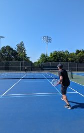 Two people playing tennis on a blue painted tennis court on a bright day