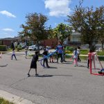 Photo: children playing on the road. They have pool noodles in their hands and a hockey net is present on the right.