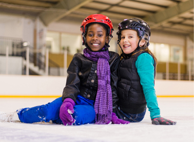 African American girl wearing a purple scarf, black coat, red helmet and blue pants sitting on the ice with a Caucasian girl wearing a teal shirt, black vest and black helmet.
