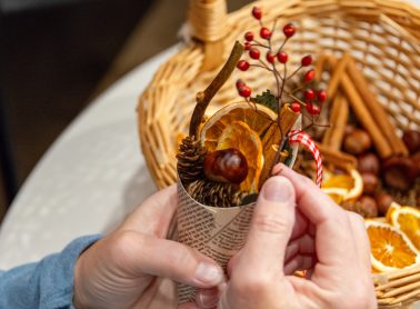 Someone holding a cone-shaped Christmas decoration with dried orange slices, cinnamon sticks, twigs, dried chestnuts, and Christmas pattern-style ribbon in a basket. Click to see full image.