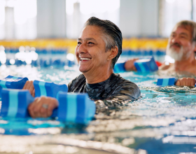 Close-up photo of a man swimming in a pool and smiling. He is holding a water dumbbell.