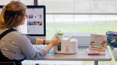 A woman is photographed sitting at a desk facing the window and using a Cricut Mug Press machine and an iMac.