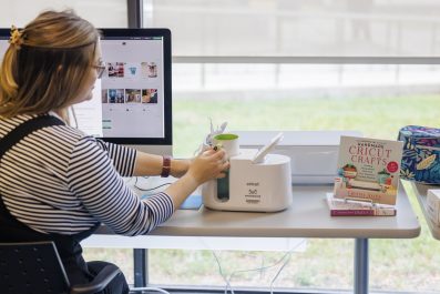 A woman is photographed sitting at a desk facing the window and using a Cricut Mug Press machine and an iMac.
