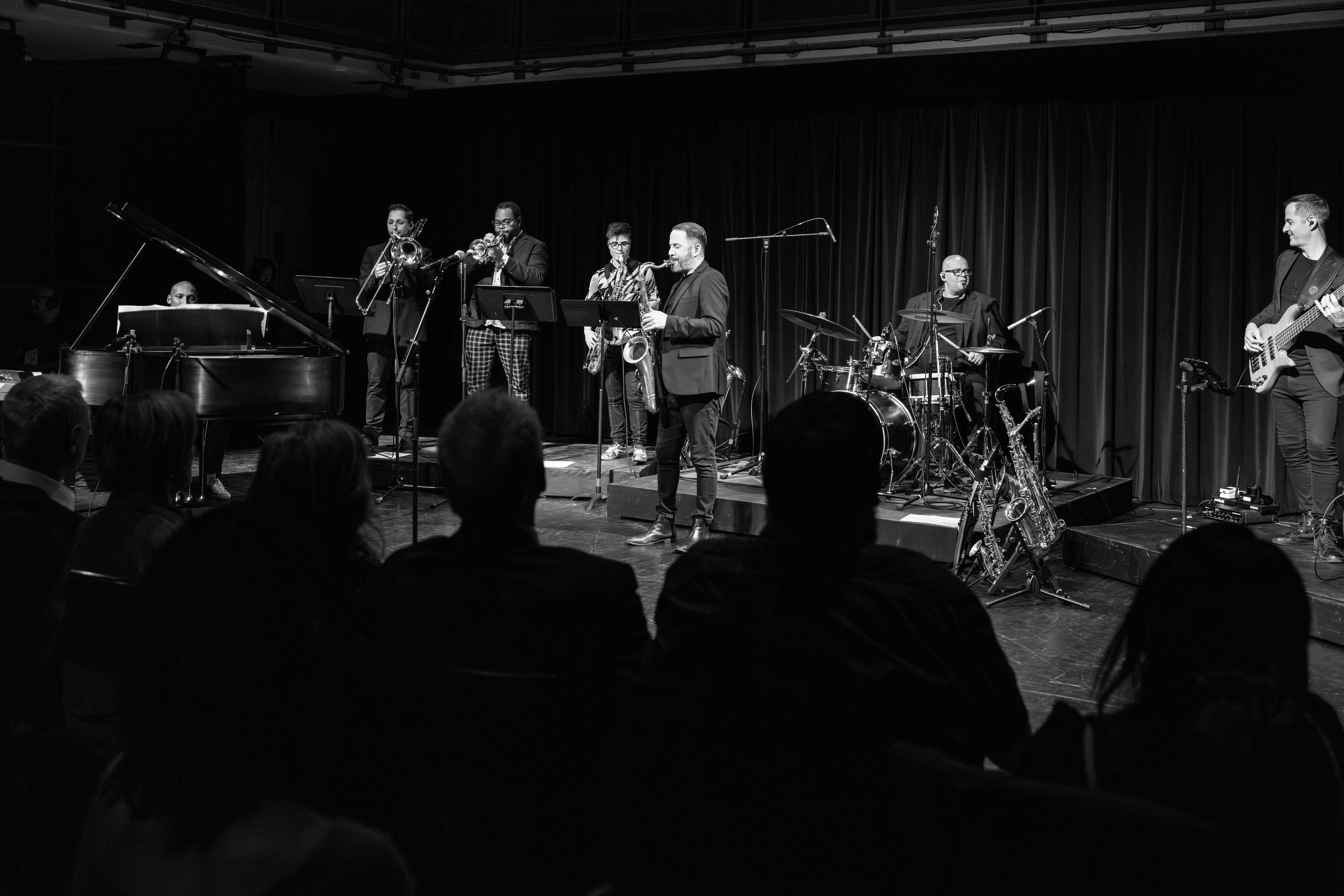 A jazz ensemble performs onstage in a dimly lit venue. Musicians play piano, brass instruments, saxophones, drums, and bass while an audience watches from their seats.