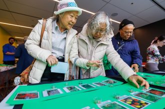 A group of participants in an indoor event space examines a table with green display boards that hold rows of photo cards and small clear containers filled with different seed types.