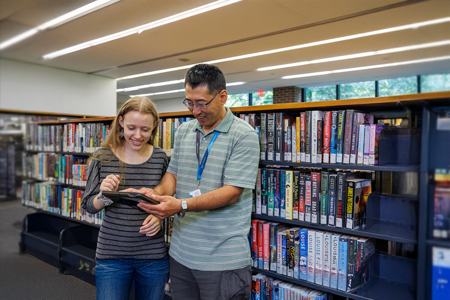 Two people using a tablet in front of library bookshelves