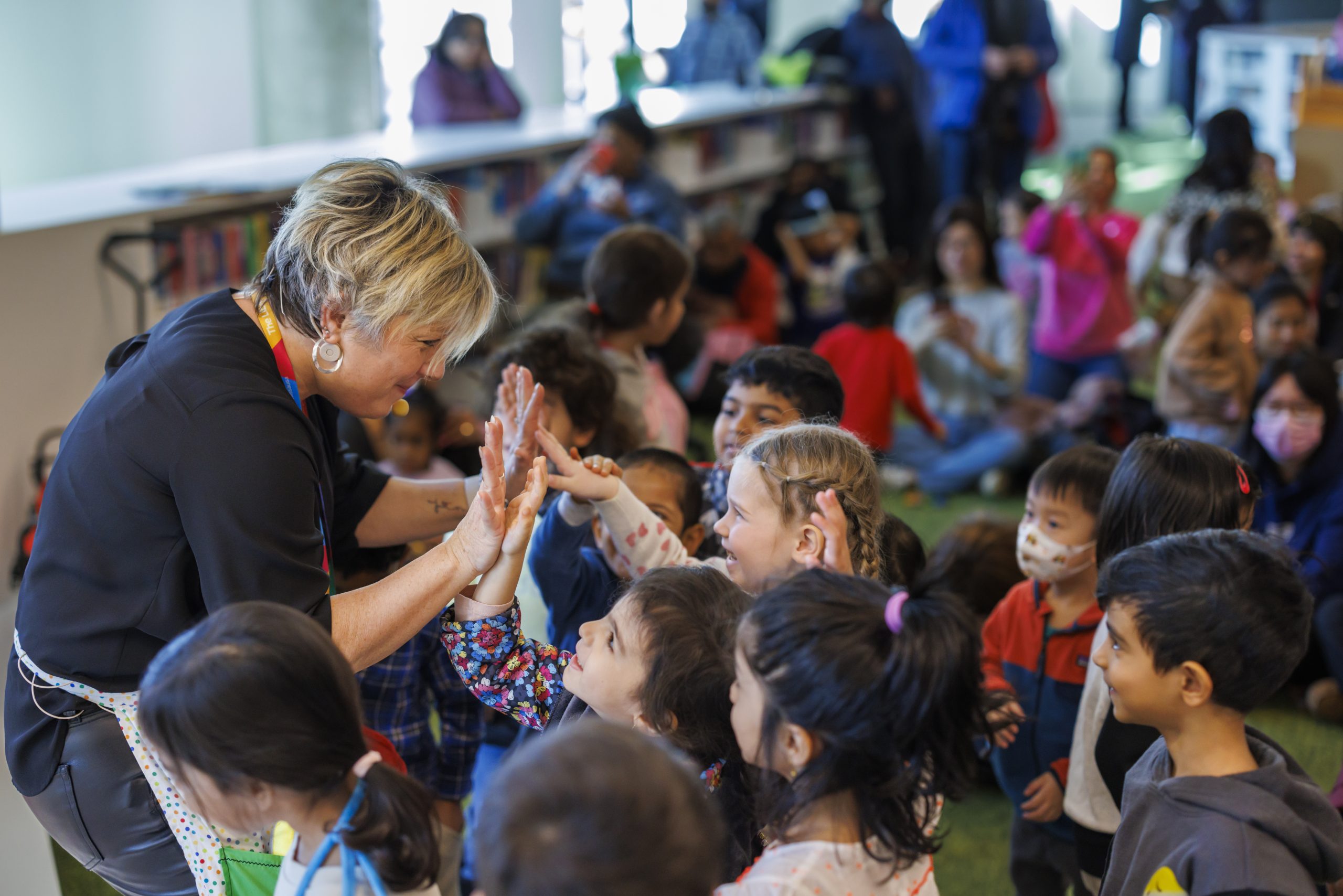 An adult interacts with young children by exchanging high-fives during a group activity inside a public library, with many children gathered around.