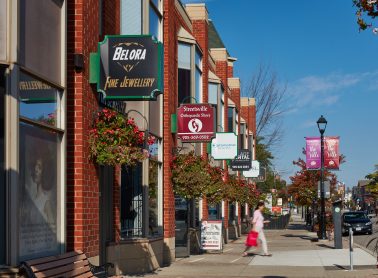 A sunny main street lined with brick storefronts and hanging flower baskets, with business signs along the sidewalk and a pedestrian walking past parked cars.