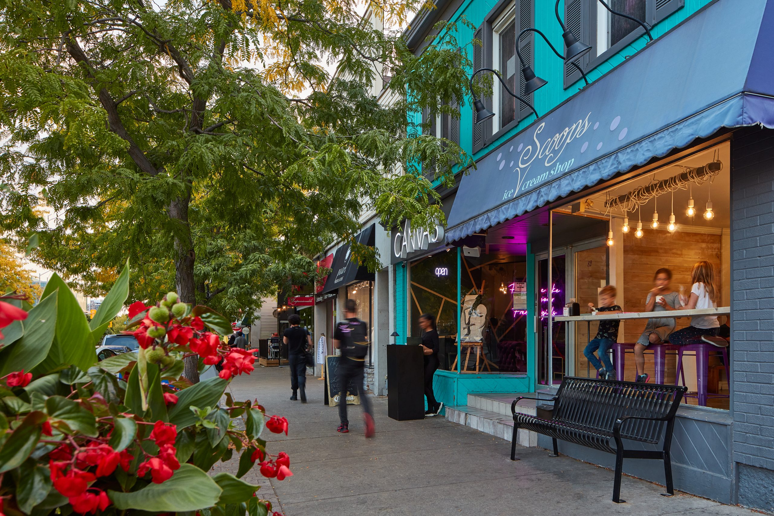 A tree‑lined sidewalk with small storefronts, including an ice cream shop with a blue awning, where pedestrians walk past and people sit at a window counter in early evening light.