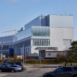 A large, modern industrial building with glass and light‑coloured exterior panels rises behind a parking lot filled with cars, under a clear blue sky.