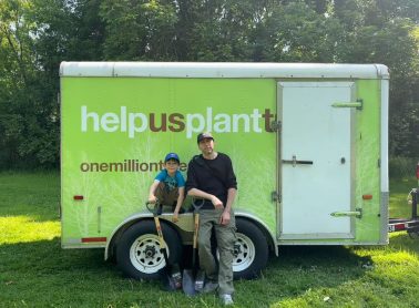 Two people holding shovels in front of a green trailer promoting ‘Help us plant’ and ‘One Million Trees’.