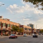 A wide view of a busy urban street with brick, multi‑storey commercial buildings, storefronts at street level, and cars driving past under a partly cloudy sky.