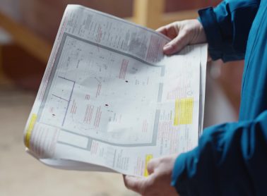 Person wearing a blue City of Mississauga jacket is in a partially framed interior space, reviewing printed construction drawings while standing on a subfloor.