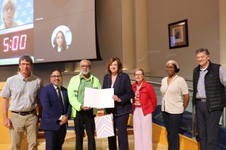 Seven people stand on a stage at Mississauga Civic Centre; two people in the centre hold an open certificate and a gift bag, with a projection screen behind them.