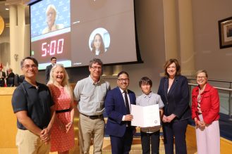 Seven people stand on a stage at Mississauga Civic Centre; one person in the centre holds an award certificate, with a projection screen visible behind them.
