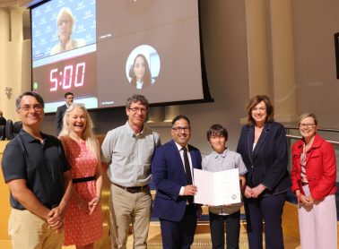 Seven people stand on a stage at Mississauga Civic Centre; two people in the centre hold an award certificate, with a projection screen visible behind them.