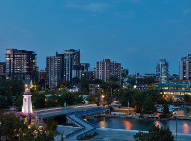 Evening waterfront view of Port Credit featuring high-rise buildings, restaurants with patios, a lighthouse, and a bridge over calm water.