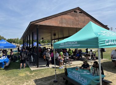 Outdoor community gathering at a park pavilion with multiple service tents and tables, including a ‘Peel Outreach – Unhoused Community Support’ booth, and people seated and standing throughout the area.