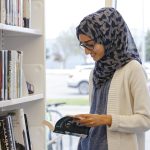 A teen library visitor stands beside a bookshelf and reads an open book. Shelves filled with books and media materials are visible in the library.