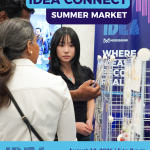 A person shows products to another person with a booth in the background.