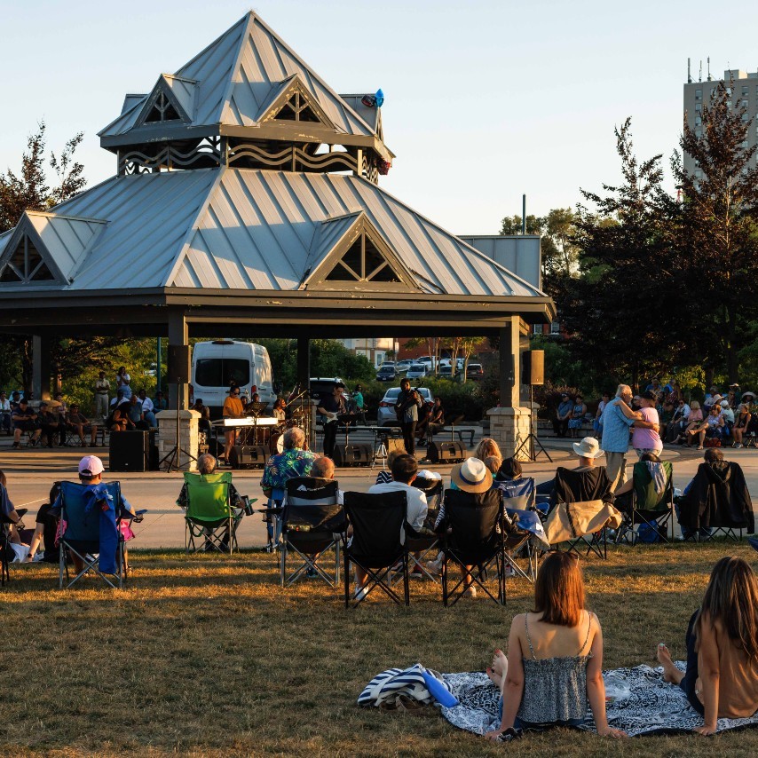 Groups of people sitting on a lawn, watching a musical performance in a park.