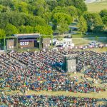 Crowd of people in an outdoor setting within trees and near a stage.