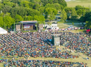 Crowd of people in an outdoor setting within trees and near a stage.