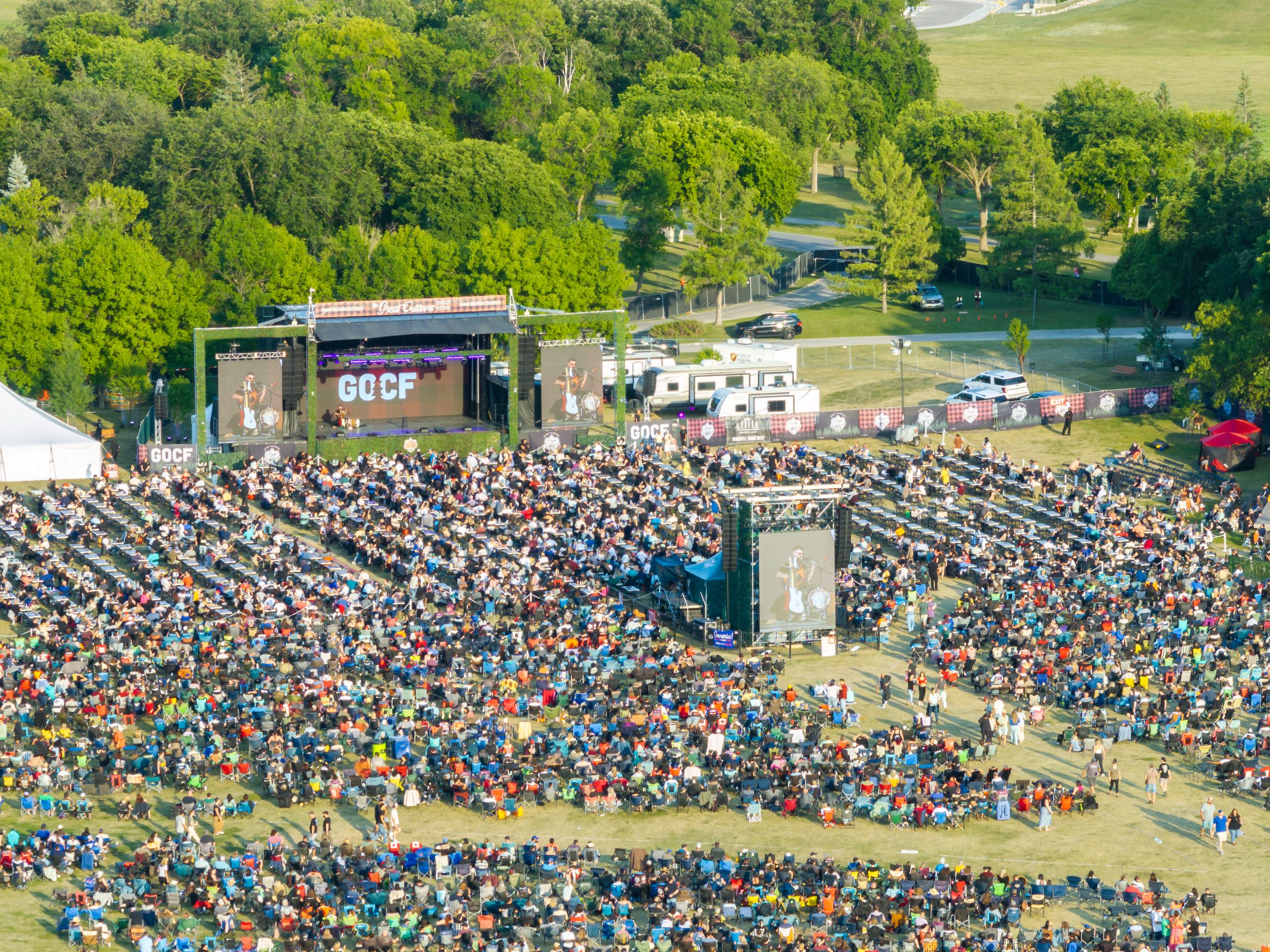 Large crowd of people sitting in front of a stage in a setting with grass and trees.