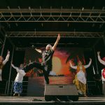 Five performers dance on an outdoor stage during a live cultural performance. One performer stands at centre stage with arms raised while four dancers in colourful traditional attire perform on either side. A large digital screen displaying a sunset scene forms the backdrop, with stage equipment and a DJ booth visible behind the performers.