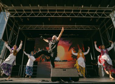 Five performers dance on an outdoor stage during a live cultural performance. One performer stands at centre stage with arms raised while four dancers in colourful traditional attire perform on either side. A large digital screen displaying a sunset scene forms the backdrop, with stage equipment and a DJ booth visible behind the performers.
