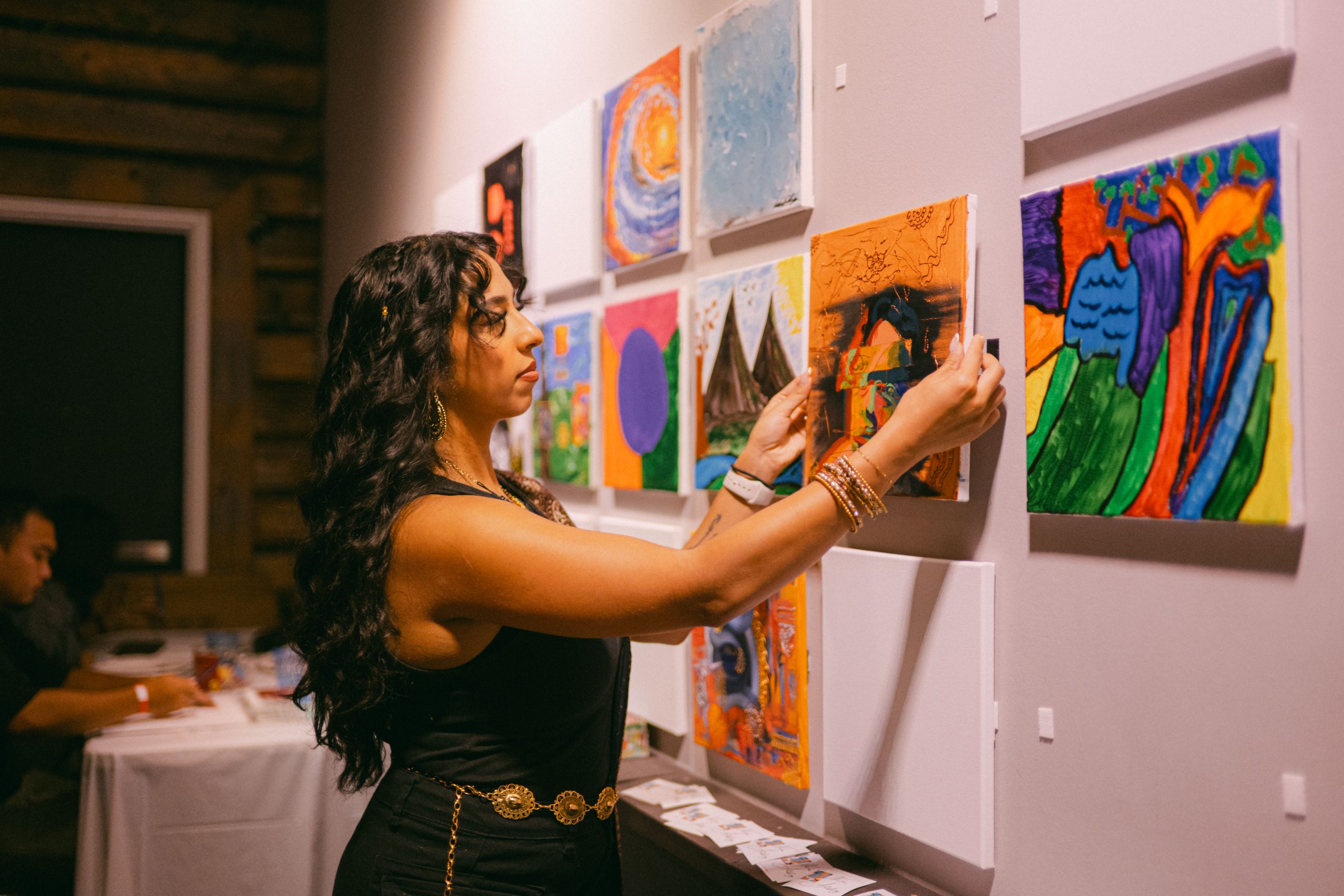 A person adjusts a colourful painting on a gallery wall displaying multiple small artworks. The indoor exhibition space features a variety of abstract and landscape-style paintings arranged in rows, with tables and event attendees visible in the background.