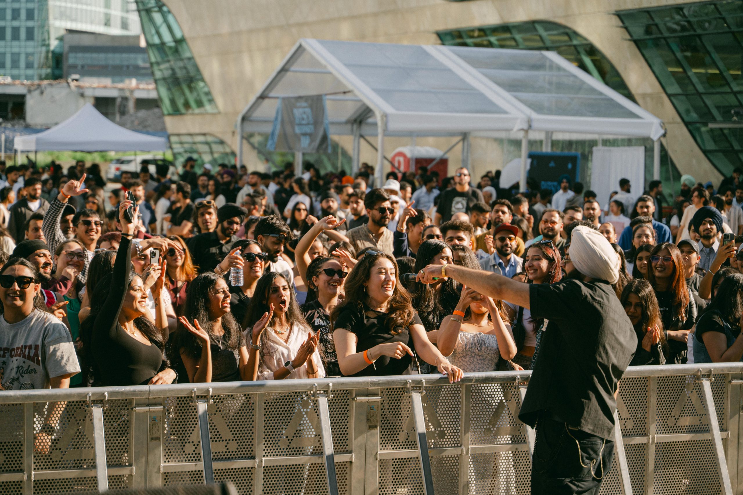A performer stands in front of a large outdoor crowd behind a metal barrier, gesturing toward attendees gathered in a plaza. Temporary event tents and a modern building with large glass windows are visible in the background. Numerous audience members are watching, holding phones and raising their hands during the daytime event.